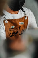 A person wearing a white shirt and a brown apron with text and a logo. The apron has metal loops and the person is also wearing a black face mask. There is a name tag on the apron.
