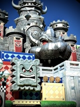 A castle with dark stone towers and aggressive medieval architectural elements, including horns and spiked decorations. In the foreground, a metallic animated face with red eyes and a fierce expression is set on a colorful blocky structure, adorned with cartoonish details like coins and patterned blocks. Brightly colored banners with an emblem add to the fantastical theme.