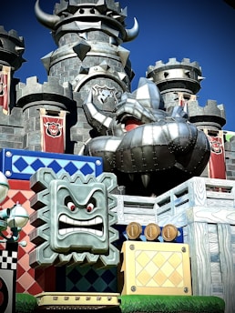 A castle with dark stone towers and aggressive medieval architectural elements, including horns and spiked decorations. In the foreground, a metallic animated face with red eyes and a fierce expression is set on a colorful blocky structure, adorned with cartoonish details like coins and patterned blocks. Brightly colored banners with an emblem add to the fantastical theme.