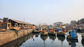 A busy harbor scene with colorful fishing boats lined up on calm water, reflecting their vivid hues. On the left, a stone dock is populated with people and various goods, including baskets and nets, beneath a roofed structure. Seagulls fly above in the slightly hazy sky, adding to the bustling yet tranquil atmosphere.