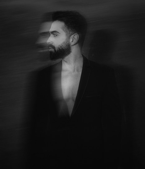 A monochrome image of a man with a beard wearing a blazer, standing against a dark background. The image has a motion blur effect, creating a ghostly duplicate of the man's face, adding a surreal and artistic quality.