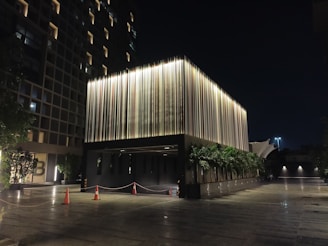 A modern building illuminated at night. The structure features vertical light strips on its facade that create a striking visual effect. There are orange traffic cones and a cordoned-off area near the entrance, with greenery and trees on both sides. The background includes another building with uniformly lit windows.