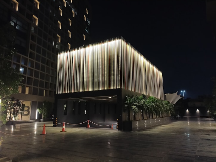 A modern building illuminated at night. The structure features vertical light strips on its facade that create a striking visual effect. There are orange traffic cones and a cordoned-off area near the entrance, with greenery and trees on both sides. The background includes another building with uniformly lit windows.