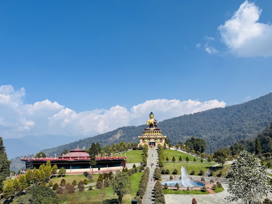 A large Buddha statue is prominently displayed on a hilltop, surrounded by manicured gardens and pathways. A fountain can be seen in the foreground, with water streaming upwards into the blue sky. The landscape features lush green hills and a partially cloudy sky, adding depth and serenity to the scene.