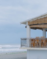 A beachfront structure with a balcony featuring wooden furniture, overlooking an overcast sky and waves crashing onto the shore. The building has a thatched roof shade and is surrounded by sandy landscape.