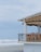 A beachfront structure with a balcony featuring wooden furniture, overlooking an overcast sky and waves crashing onto the shore. The building has a thatched roof shade and is surrounded by sandy landscape.