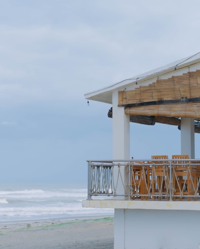A beachfront structure with a balcony featuring wooden furniture, overlooking an overcast sky and waves crashing onto the shore. The building has a thatched roof shade and is surrounded by sandy landscape.