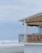 A beachfront structure with a balcony featuring wooden furniture, overlooking an overcast sky and waves crashing onto the shore. The building has a thatched roof shade and is surrounded by sandy landscape.