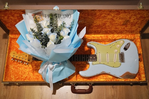 A light blue electric guitar is placed in an open guitar case with a vibrant orange interior. A bouquet of blue and white flowers, wrapped in blue paper and accompanied by a card, is resting on top of the guitar.
