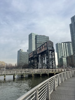 A waterfront scene with a large industrial structure featuring the words 'Long Island' prominently displayed. Tall modern buildings are situated in the background under a cloudy sky. A boardwalk with metal railings runs parallel to the water.
