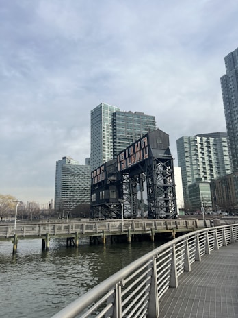 A waterfront scene with a large industrial structure featuring the words 'Long Island' prominently displayed. Tall modern buildings are situated in the background under a cloudy sky. A boardwalk with metal railings runs parallel to the water.