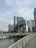 A waterfront scene with a large industrial structure featuring the words 'Long Island' prominently displayed. Tall modern buildings are situated in the background under a cloudy sky. A boardwalk with metal railings runs parallel to the water.