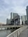 A waterfront scene with a large industrial structure featuring the words 'Long Island' prominently displayed. Tall modern buildings are situated in the background under a cloudy sky. A boardwalk with metal railings runs parallel to the water.