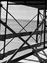 Black and white image of scaffolding with wooden planks and metal bars intersecting in various angles. A netting or mesh covers the background, and there is a view of a body of water beyond the structure.