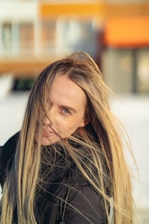 A person with long blonde hair partially covering their face, standing outdoors with a soft focus background. The person is wearing a black jacket and has an expression of calmness and introspection.