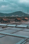 Several rustic wooden huts with rusted metal roofs are situated near rectangular water pools, surrounded by a natural landscape with hills in the background. Power lines and pylons are visible against the cloudy sky, creating an industrial contrast with the rural setting.