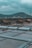 Several rustic wooden huts with rusted metal roofs are situated near rectangular water pools, surrounded by a natural landscape with hills in the background. Power lines and pylons are visible against the cloudy sky, creating an industrial contrast with the rural setting.