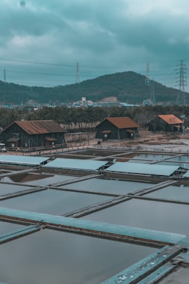 Several rustic wooden huts with rusted metal roofs are situated near rectangular water pools, surrounded by a natural landscape with hills in the background. Power lines and pylons are visible against the cloudy sky, creating an industrial contrast with the rural setting.