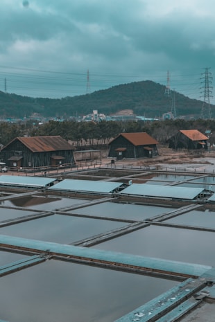Several rustic wooden huts with rusted metal roofs are situated near rectangular water pools, surrounded by a natural landscape with hills in the background. Power lines and pylons are visible against the cloudy sky, creating an industrial contrast with the rural setting.