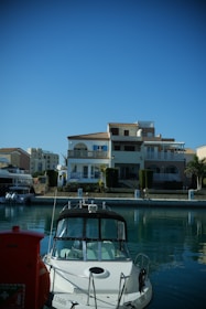 A modern, multi-story waterfront house is situated by a calm body of water, with a white motorboat docked in front of it. The house features large windows, balconies, and a light color palette with some greenery and shrubs in the surrounding area.