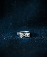 A silver ring featuring a rectangular shape with geometric designs and a prominent oval gemstone. The background is textured with a dark blue color, highlighting the ring.