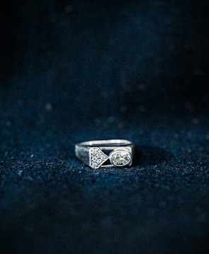 A silver ring featuring a rectangular shape with geometric designs and a prominent oval gemstone. The background is textured with a dark blue color, highlighting the ring.