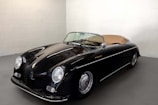 A sleek vintage convertible car with a glossy black exterior is parked in a well-lit, minimalistic room. The car features distinctive round headlights and a tan convertible top, highlighting its classic design.