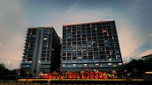 Two high-rise buildings with illuminated windows and signage are visible. The sky is in a dusky state with shades of blue and gray. A brightly lit sign reading 'HELLO AHMEDABAD' is displayed at the foreground, indicating a welcoming message.
