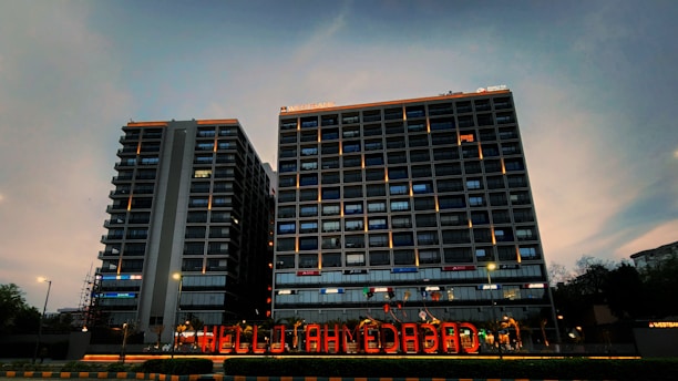 Two high-rise buildings with illuminated windows and signage are visible. The sky is in a dusky state with shades of blue and gray. A brightly lit sign reading 'HELLO AHMEDABAD' is displayed at the foreground, indicating a welcoming message.