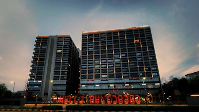 Two high-rise buildings with illuminated windows and signage are visible. The sky is in a dusky state with shades of blue and gray. A brightly lit sign reading 'HELLO AHMEDABAD' is displayed at the foreground, indicating a welcoming message.