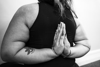 A person is performing a reverse prayer pose, known in yoga as Paschim Namaskarasana, with their hands joined behind their back. The image is in black and white, highlighting the texture of the skin and the tattoo on the individual's arm.