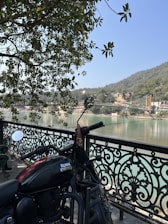 A Royal Enfield motorcycle is parked next to an ornate black metal railing, with a view of a calm river and a distant hillside. A suspension bridge spans the river in the background, and the area is surrounded by lush greenery. Sunlight filters through the leaves of a tree overhead.