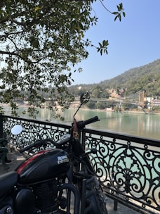 A Royal Enfield motorcycle is parked next to an ornate black metal railing, with a view of a calm river and a distant hillside. A suspension bridge spans the river in the background, and the area is surrounded by lush greenery. Sunlight filters through the leaves of a tree overhead.