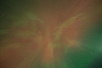 A night sky illuminated by a vibrant aurora, displaying swirling colors of green and red intermingling against a backdrop of scattered stars. The aurora creates an ethereal, otherworldly pattern that seems to flow across the sky.