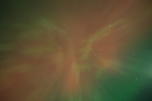 A night sky illuminated by a vibrant aurora, displaying swirling colors of green and red intermingling against a backdrop of scattered stars. The aurora creates an ethereal, otherworldly pattern that seems to flow across the sky.