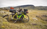 A brightly colored bicycle is fully loaded with camping gear, including bags on the frame, handlebars, and rear wheel. It rests against a large rock in a grassy, open landscape with distant hills under an overcast sky.