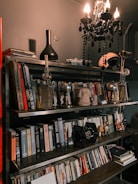 A bookcase is filled with a variety of books arranged on multiple shelves. Above the shelves, a black chandelier with lit bulbs hangs, casting light on the surrounding area. There are decorative items on the shelves, including a black vase, a helmet, and various figurines and models like a TIE fighter and robots.