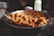 A large metal pot filled with a generous serving of meatballs coated in a rich tomato-based sauce. A metal serving spoon rests against the edge of the pot. The setting appears to be a food stall or market, indicated by the presence of a plastic condiment bottle and an obscured background.