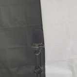 A microphone mounted on a stand is set against a background divided into a black section on the left and a white section on the right. The microphone has a black foam cover, and the stand appears to be adjustable.