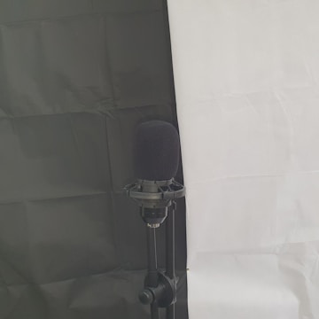A microphone mounted on a stand is set against a background divided into a black section on the left and a white section on the right. The microphone has a black foam cover, and the stand appears to be adjustable.