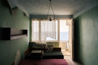 A small, dimly lit room with light green floral wallpaper, featuring a large window with partially drawn, sheer curtains that allow a view of the sea. The room contains a bed with a pink mattress, a small table, a dark couch, a wall-mounted shelf, and an old radiator. A glass chandelier hangs from the ceiling, and the door next to the window is open, leading to a narrow balcony.