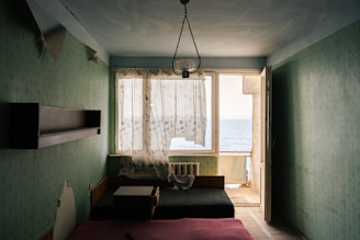 A small, dimly lit room with light green floral wallpaper, featuring a large window with partially drawn, sheer curtains that allow a view of the sea. The room contains a bed with a pink mattress, a small table, a dark couch, a wall-mounted shelf, and an old radiator. A glass chandelier hangs from the ceiling, and the door next to the window is open, leading to a narrow balcony.