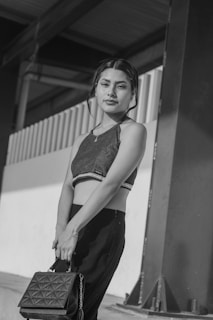 A woman stands confidently while holding a handbag, positioned against a backdrop of industrial elements such as beams and corrugated metal. The image is in black and white, emphasizing contrast and textures.