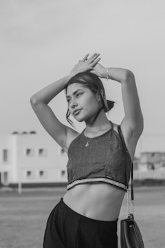 A monochrome photo featuring a young woman outdoors with her hands posed above her head. She is wearing a sleeveless crop top, black pants, and a necklace. There is a modern building in the background with windows and a grassy field in the foreground.