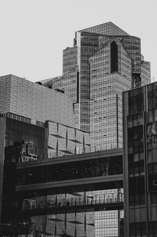 A modern cityscape featuring tall, geometric buildings made mostly of glass, displaying reflections and providing a complex architectural pattern. A skybridge connects two structures, and there are visible silhouettes of people on the bridge.