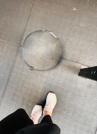 A gym setting features a medicine ball resting on a textured rubber floor. A person wearing light-colored athletic shoes stands nearby, suggesting a workout environment.