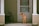 A brown dog with a green collar sits on the porch, looking to the side. The porch has light tan siding and white columns with a closed white door behind the dog. A couple of stacked chairs are visible to the left side of the porch.
