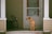A brown dog with a green collar sits on the porch, looking to the side. The porch has light tan siding and white columns with a closed white door behind the dog. A couple of stacked chairs are visible to the left side of the porch.