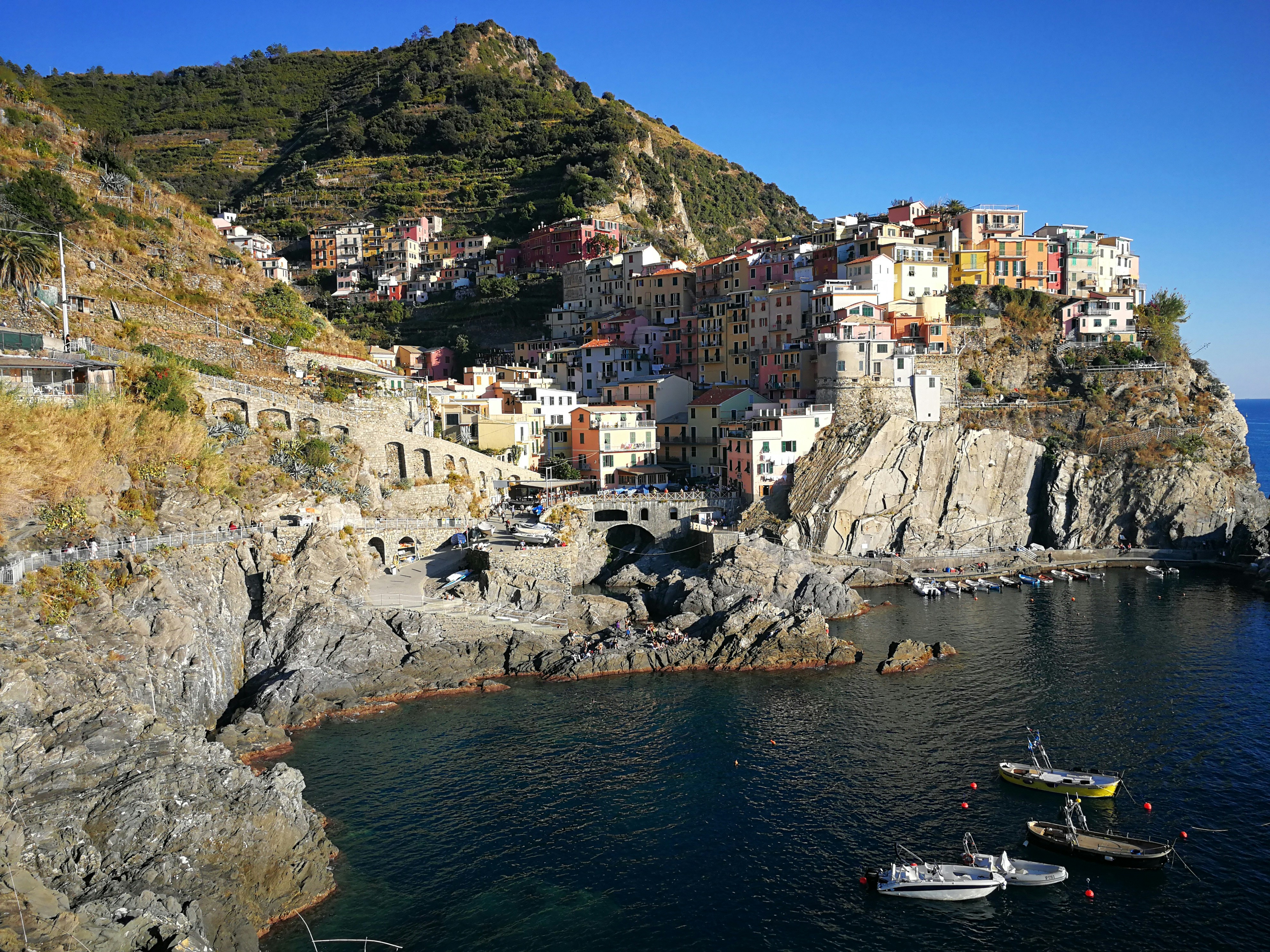 Colorful fishing boats moored in a Cinque Terre harbor with pastel-painted houses stacked on dramatic cliffside above the Mediterranean