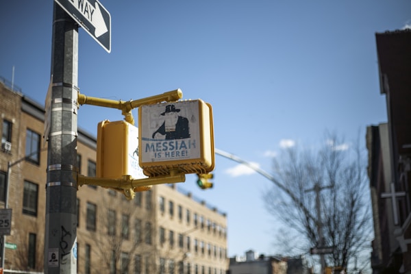 A weathered street sign with a graphic of a person wearing a hat and the phrase 'Messiah is Here!' is mounted on a yellow pedestrian signal. It is attached to a pole that also holds a one-way sign. The background features a cityscape with brick buildings and a tree, and the sky is clear and blue.